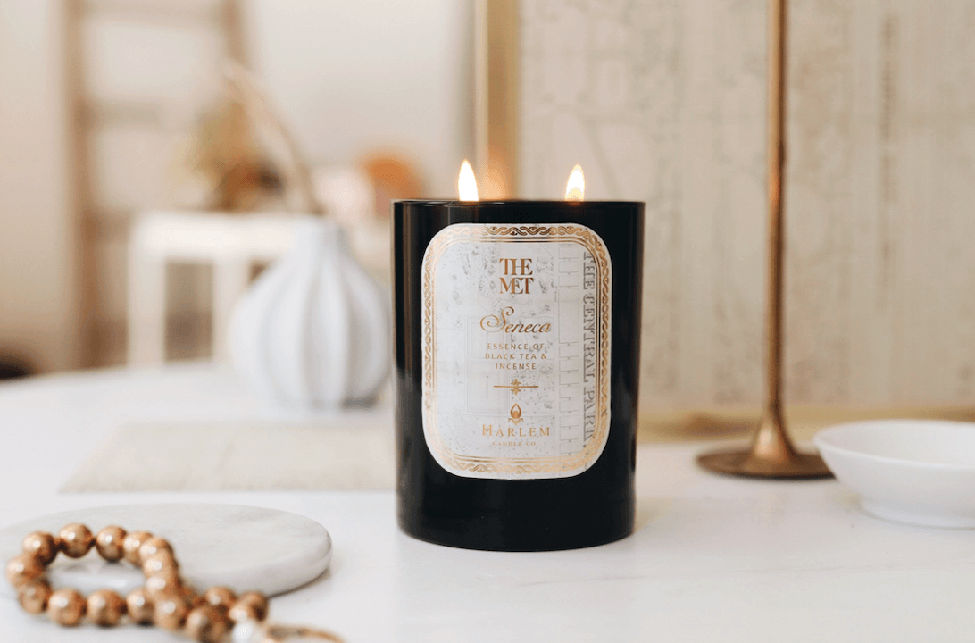 Gorgeous lifestyle image of the lit Met Seneca 12 oz candle sitting on a white table with gold jewelry and gold candle holders.
