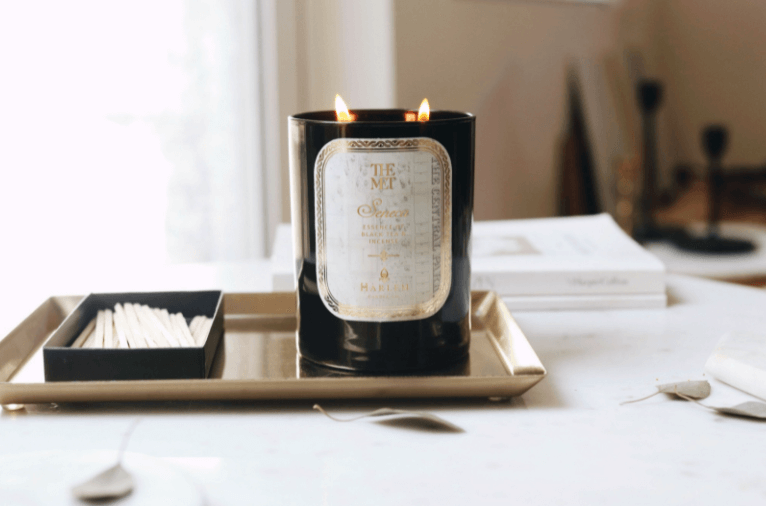 Gorgeous lifestyle image of the Met Seneca 12 oz candle sitting on a gold tray next to matches, with books and leaves on the table next to it.