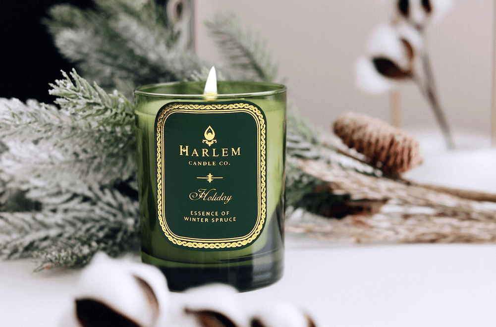 A lifestyle image of our Green Holiday Candle sitting on a table with winter spruce leaves surrounding the candle, with pinecones.