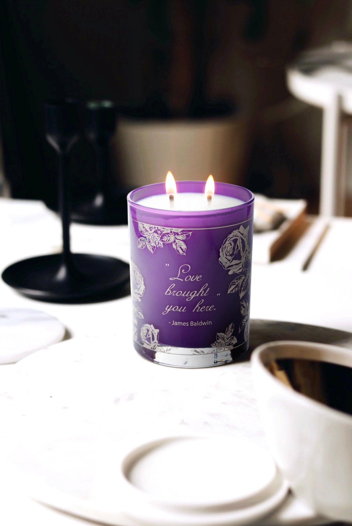 Limited edition purple and silver Love candle.  It says "Love Brought You Here" - James Baldwin. This photo was taken in a lifestyle setting with non-descript items in the background