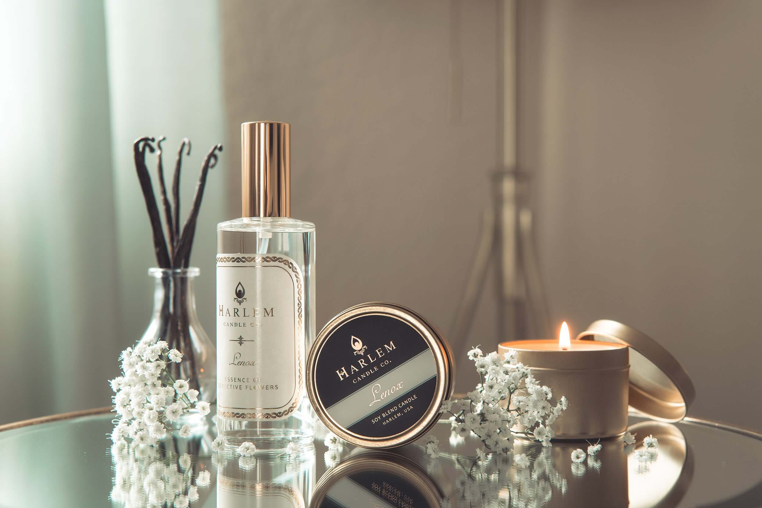 This lifestyle photo features the Lenox Room Spray and the Lenox 4 oz travel candle  as well as a lit travel tin candle and flowers with a beautiful base on a mirrored side table.